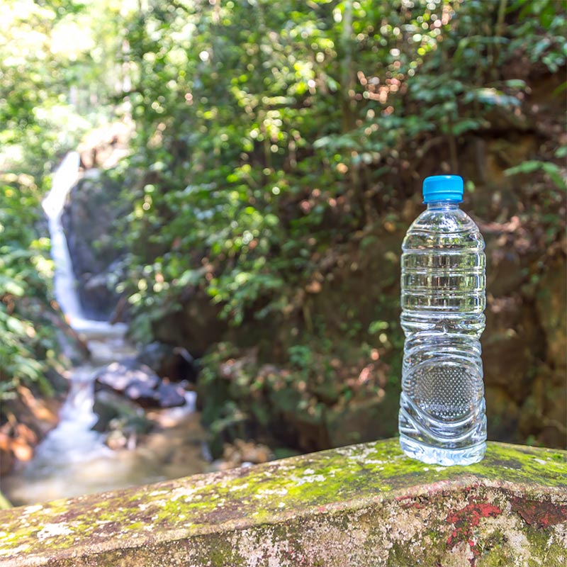 Fotografía de una botella de agua sobre una estructura de piedra con una cascada y naturaleza de fondo